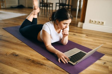 Indoor image of beautiful young woman with fit muscular body lying on her stomach on yoga mat surfing internet via laptop after home training. Barefooted girl watching online fitness video Indoor image of beautiful young woman with fit muscular body lying on her stomach on yoga mat surfing internet via laptop after home training. Barefooted girl watching online fitness video