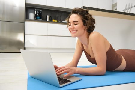 Portrait of beautiful sportswoman, fitness girl watching videos on laptop during workout, follow online pilates videos while exercise at home, wearing activewear in kitchen Portrait of beautiful sportswoman, fitness girl watching videos on laptop during workout, follow online pilates videos while exercise at home, wearing activewear in kitchen.